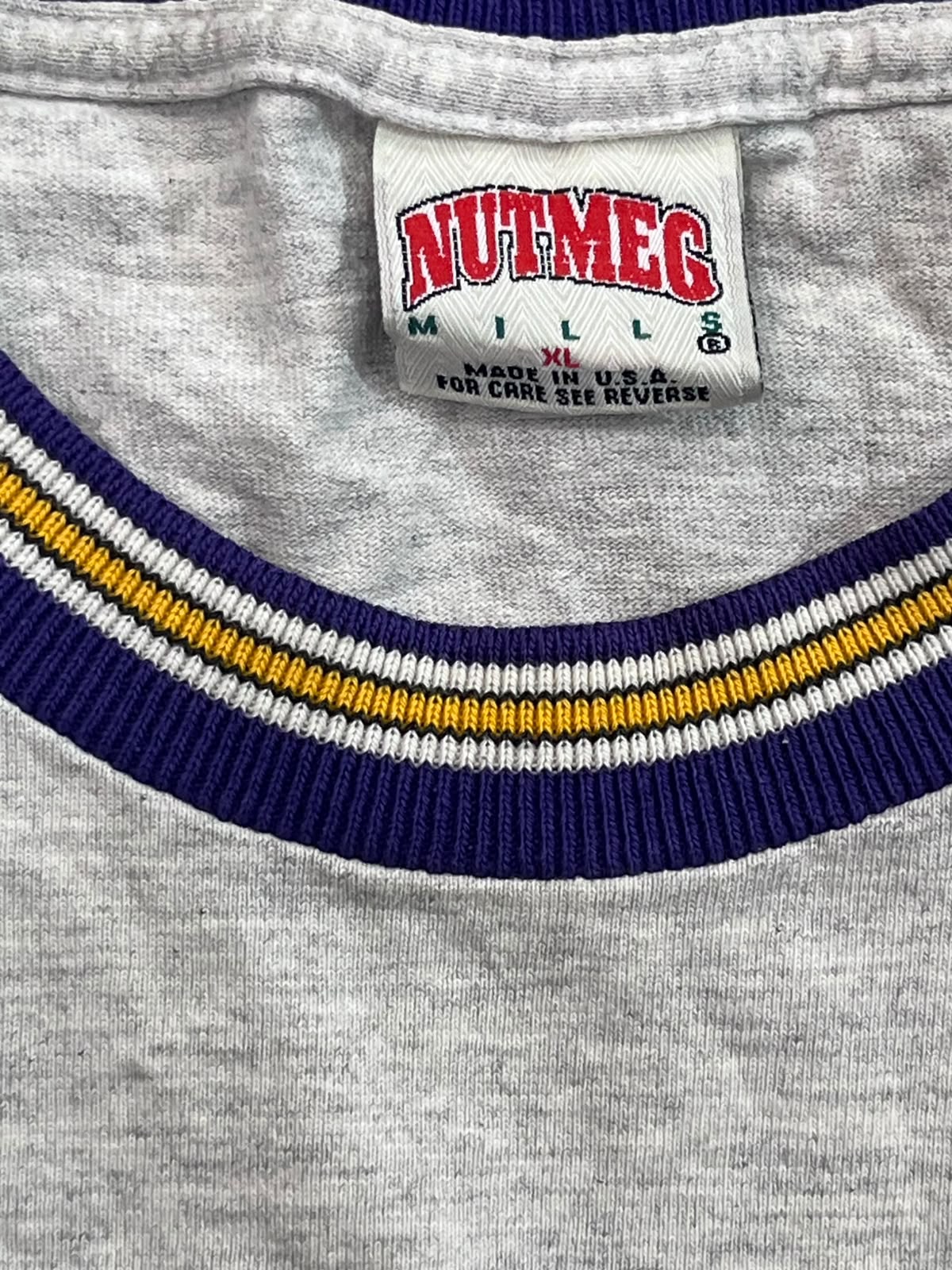 Close-up of a heather gray crewneck shirt with a navy blue and yellow striped ribbed collar. The inner neck label shows the brand 'Nutmeg Mills' in red and green text.