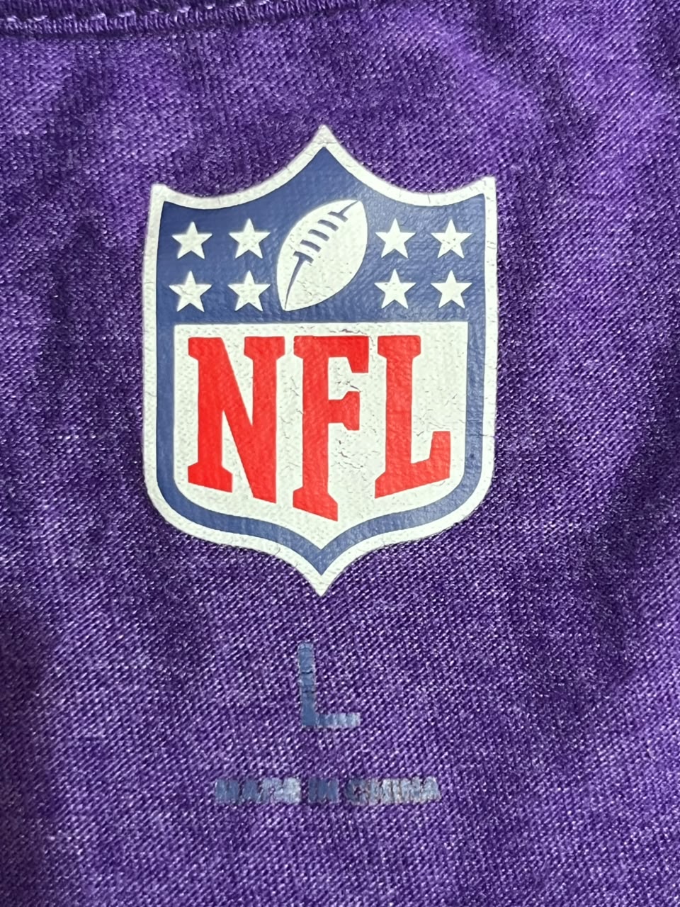 Close-up of a purple textured fabric garment featuring a large NFL shield logo on the chest and a faded size 'L' graphic below.