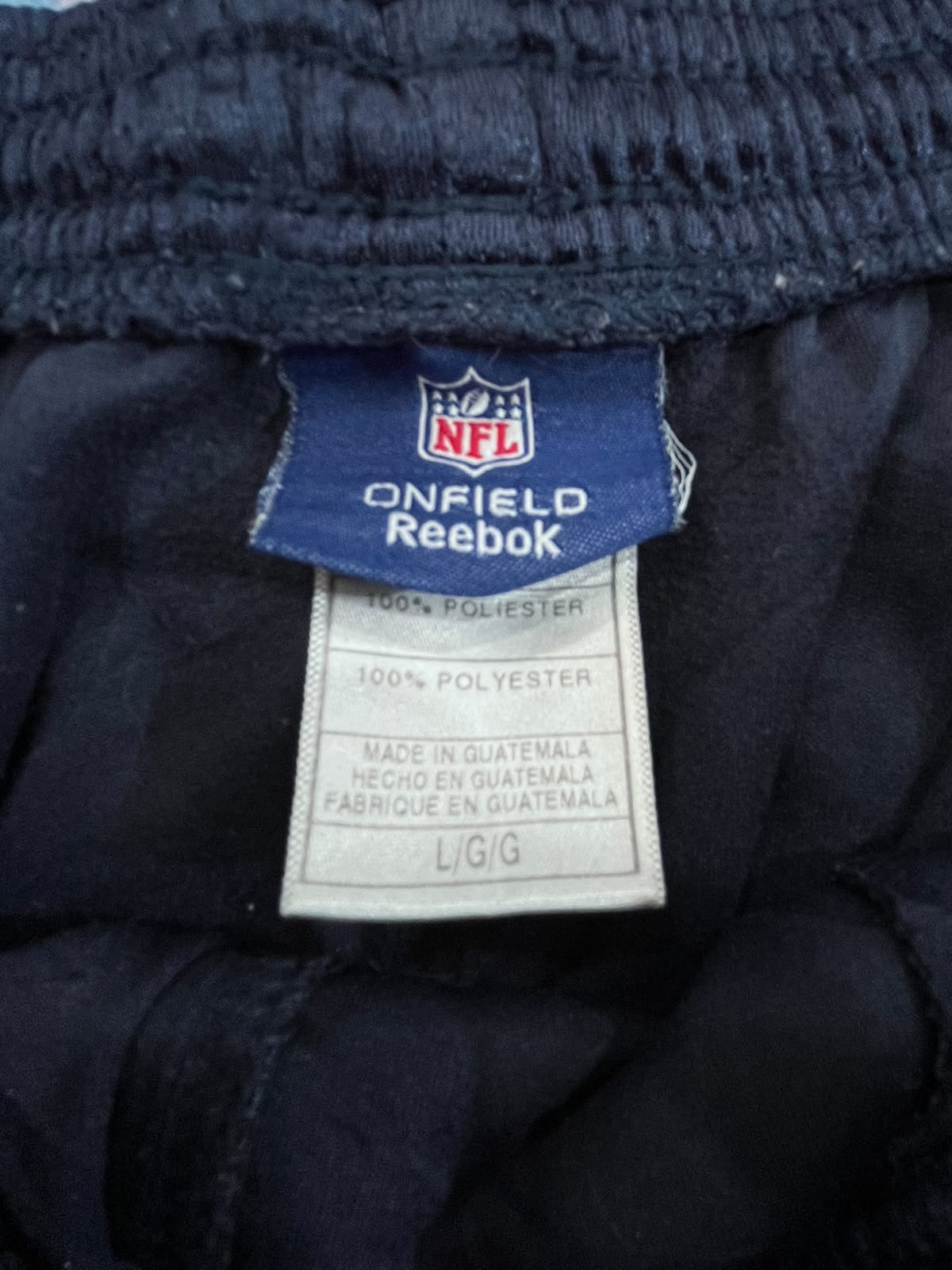 Close-up of the inner neck label of dark navy blue Reebok NFL Onfield apparel, showing the NFL shield logo and Reebok branding on a blue tag with a white size tag below.