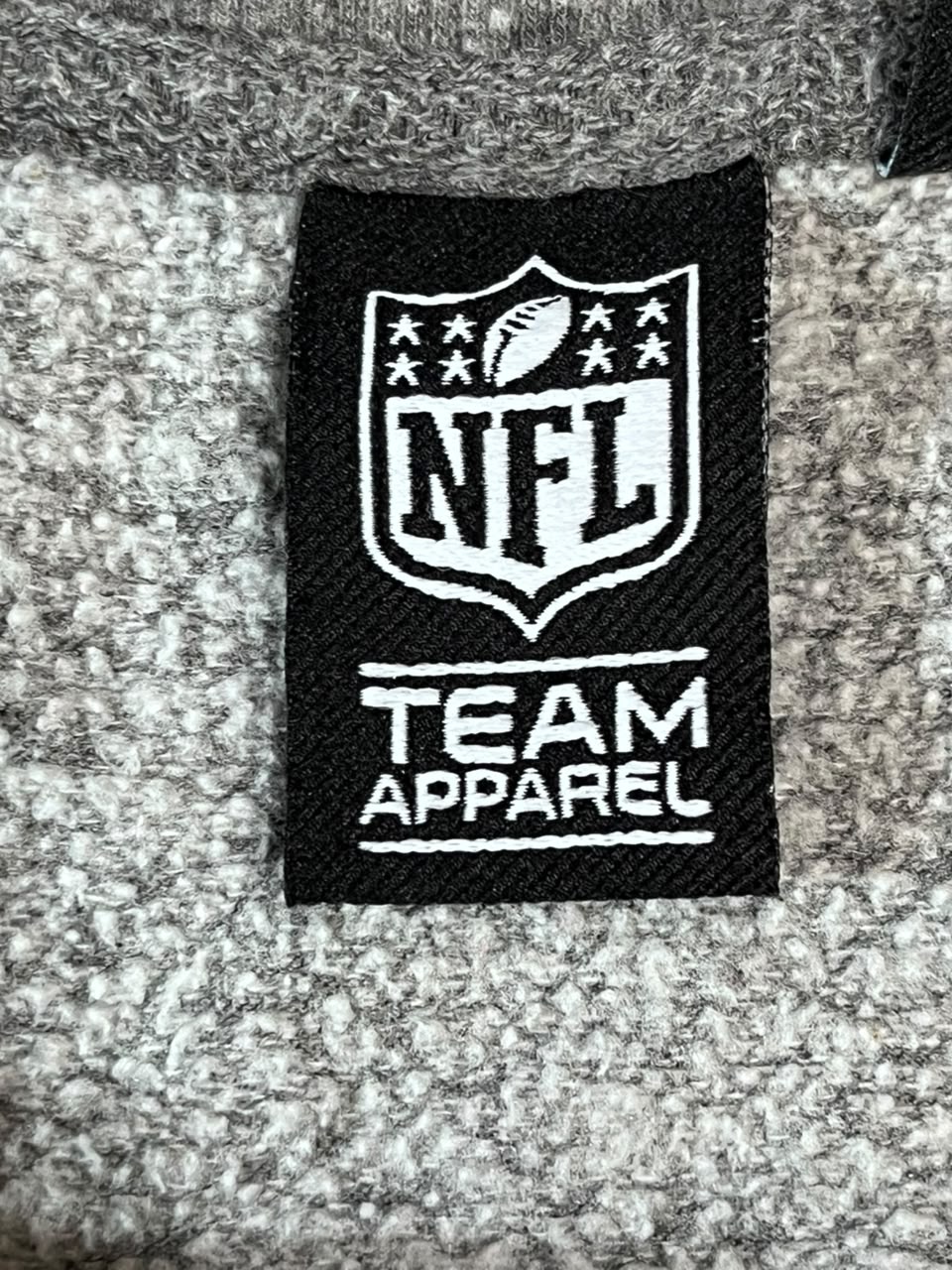 Close-up of a black NFL Team Apparel label sewn onto a gray heathered knit fabric.