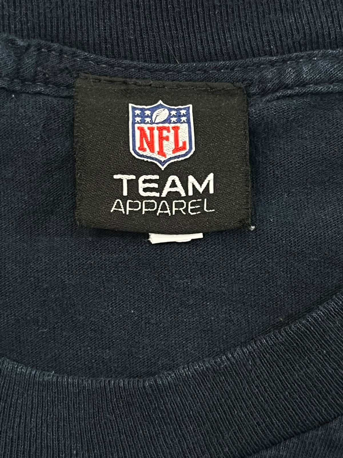 Close-up of a black neck label sewn onto a dark navy ribbed knit garment, featuring the NFL shield logo and the text 'NFL TEAM APPAREL'.