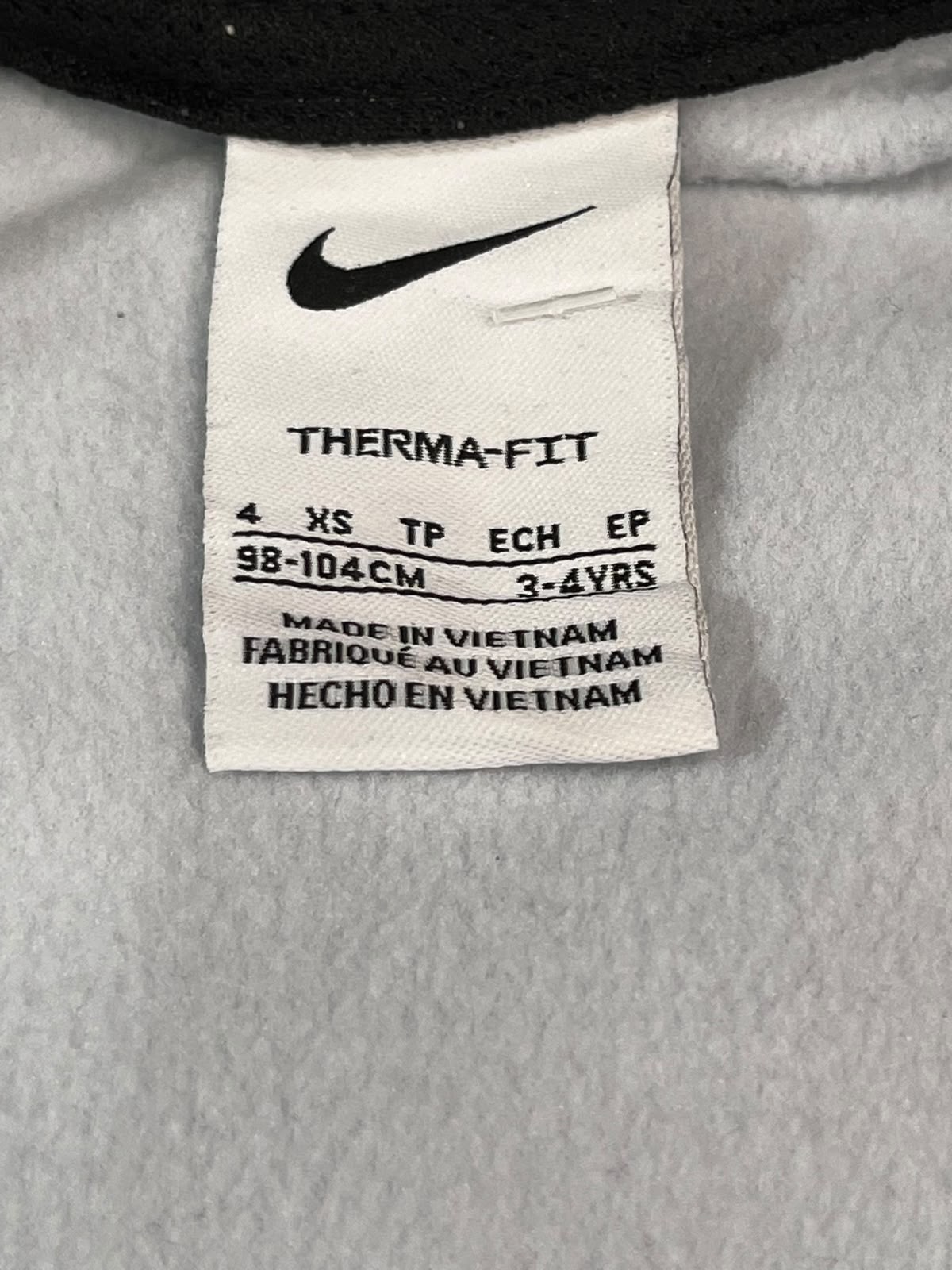 Close-up of a Nike THERMA-FIT garment interior label sewn onto light gray fabric, showing the black Nike swoosh and size information.