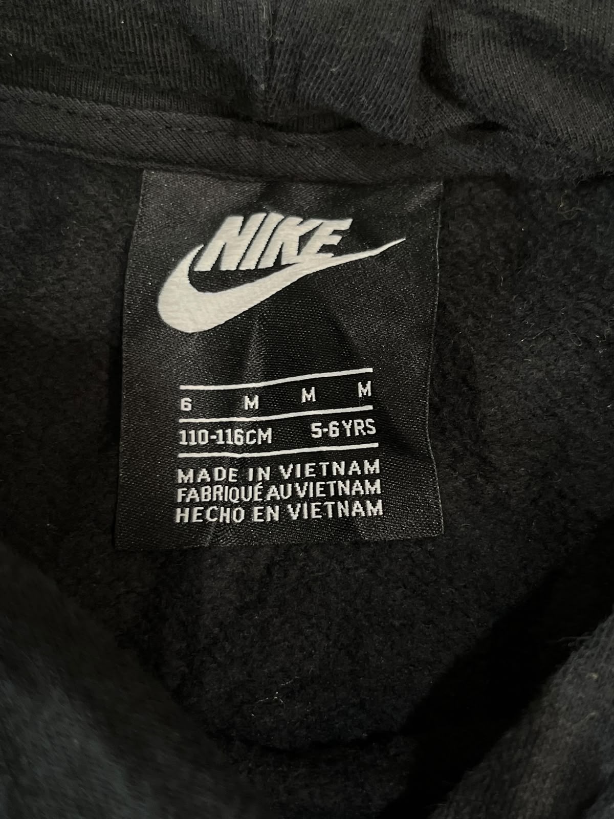 Close-up of a black Nike garment interior label showing the Nike swoosh, size information for 6 M / 110-116cm / 5-6YRS, and 'Made in Vietnam' text.