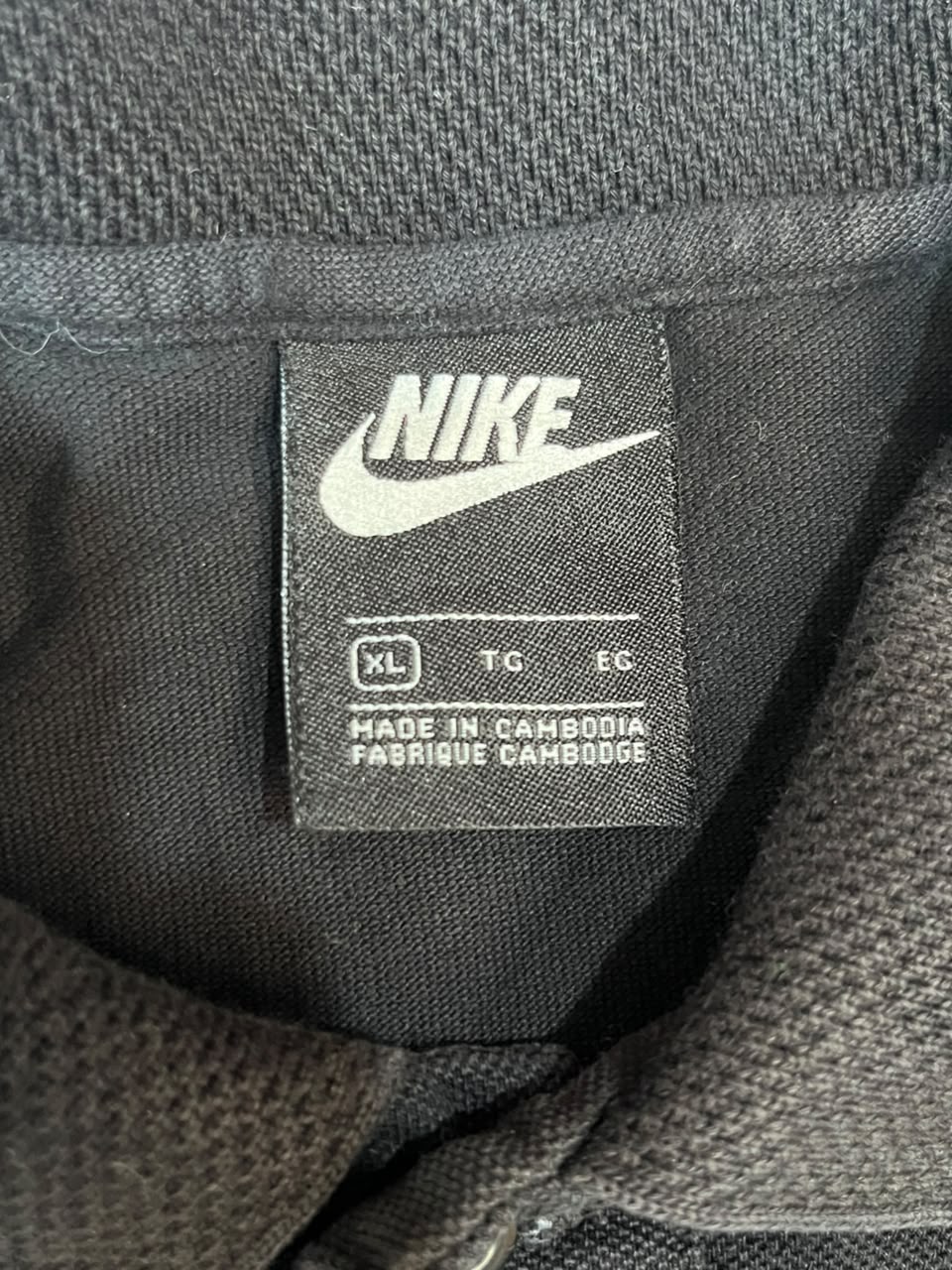 Close-up of a Nike size tag on a dark gray textured fabric garment, showing the white Nike swoosh and text including 'XL', 'T/C', 'EC', and 'MADE IN CAMBODIA'.