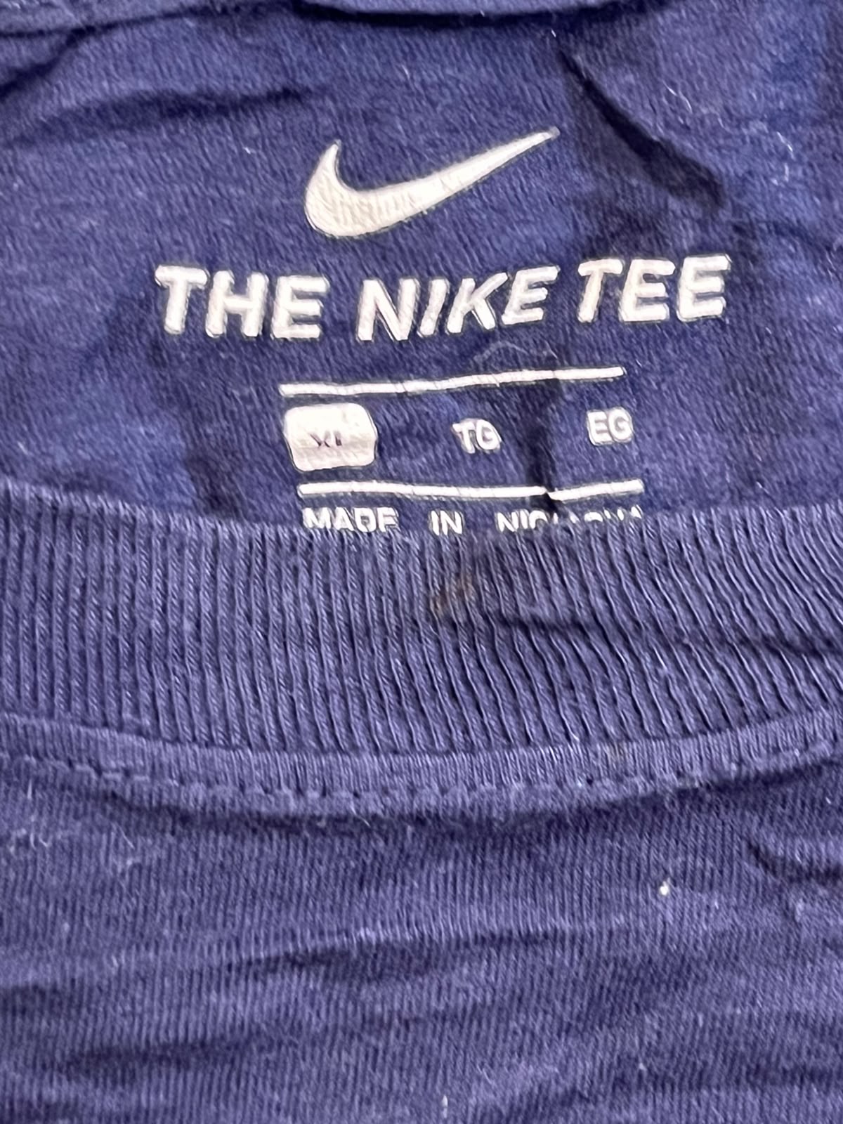 Close-up of a navy blue Nike t-shirt showing the inner neck label with a white Nike swoosh and 'THE NIKE TEE' text.