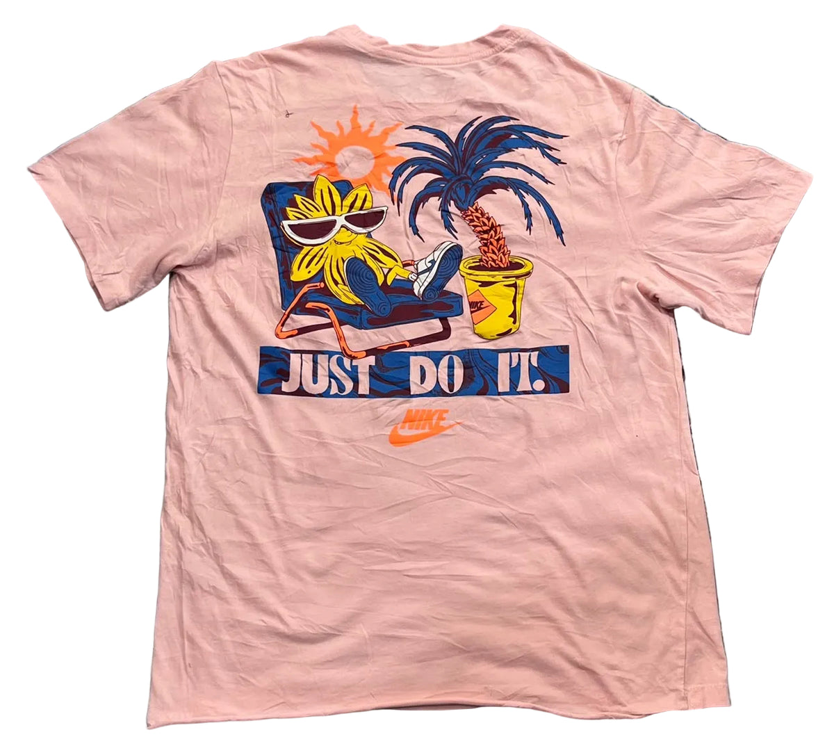A light pink short-sleeve t-shirt with a colorful graphic of a cartoon character relaxing under a palm tree with the text 'JUST DO IT.' and a small Nike swoosh, laid flat on a blue and purple shag rug.