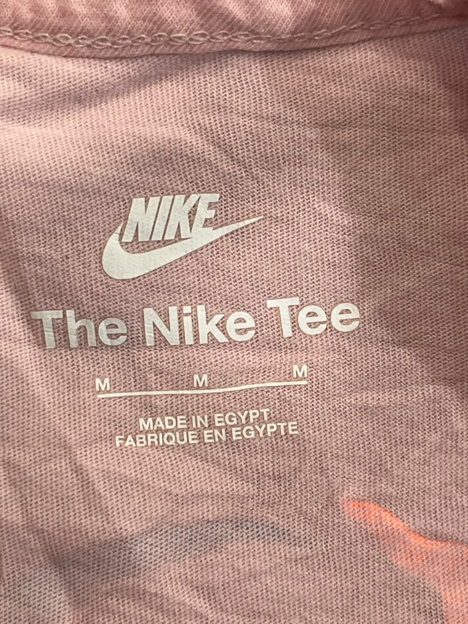 Close-up of a pink Nike t-shirt fabric with a white printed Nike logo, 'The Nike Tee' text, and 'MADE IN EGYPT FABRIQUE EN EGYPTE' on the chest.