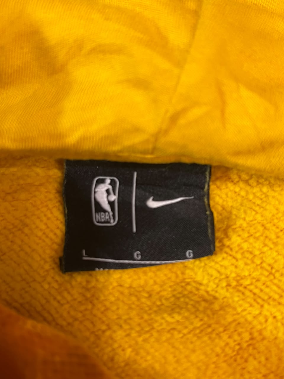 Close-up of a black Nike NBA size tag sewn onto a bright yellow-orange textured fabric, likely a sweatshirt or jacket.