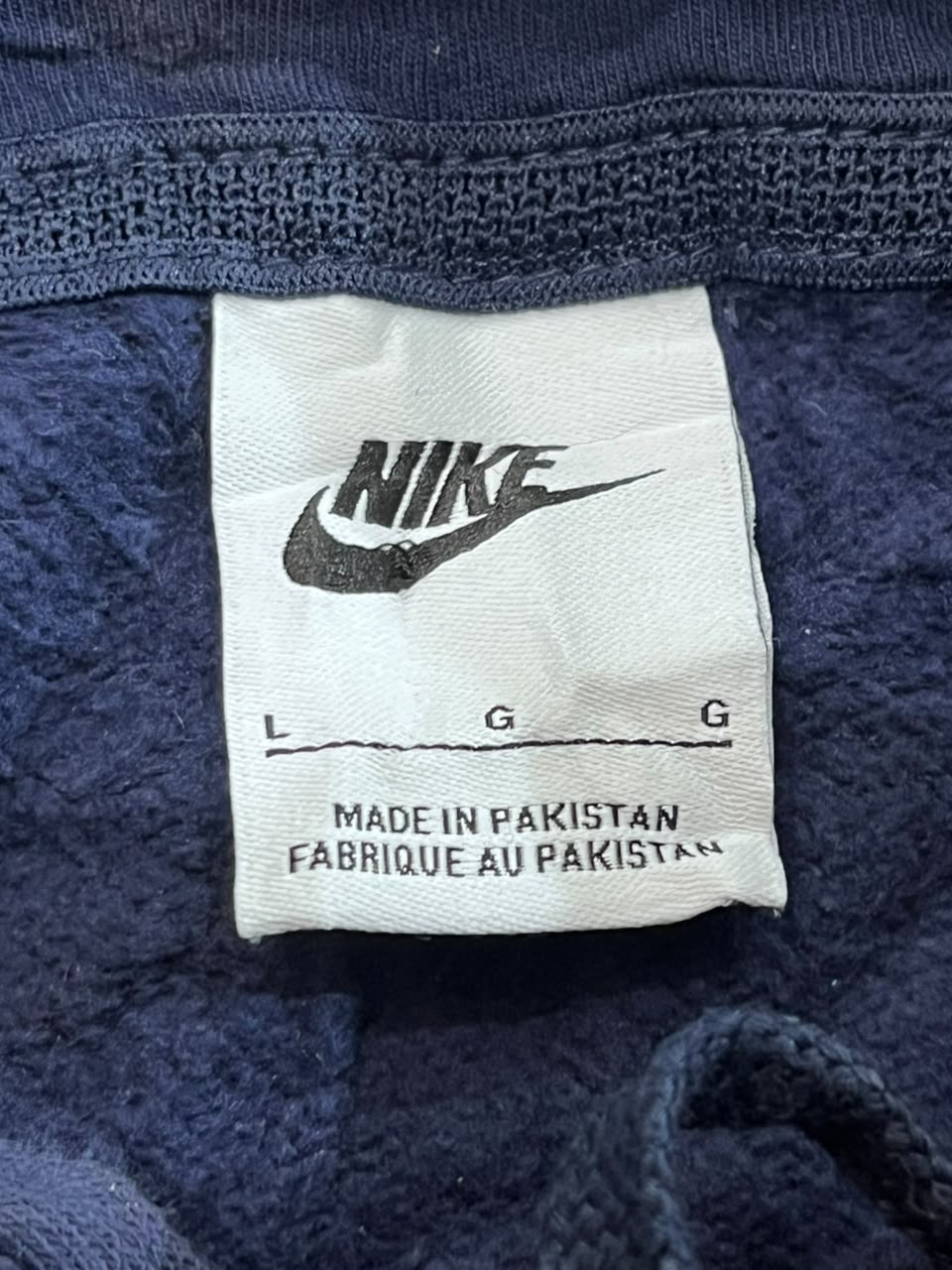 Close-up of a white Nike size tag with black printed logo and text reading 'MADE IN PAKISTAN' on a dark navy textured fabric background.