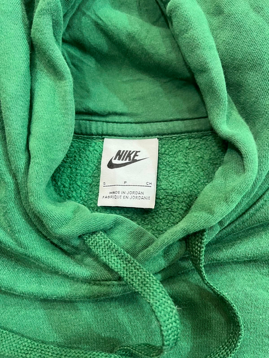 Close-up of a green Nike hoodie showing the inner neck label with the Nike logo and 'MADE IN JORDAN' text, along with green drawstrings and hood fabric.