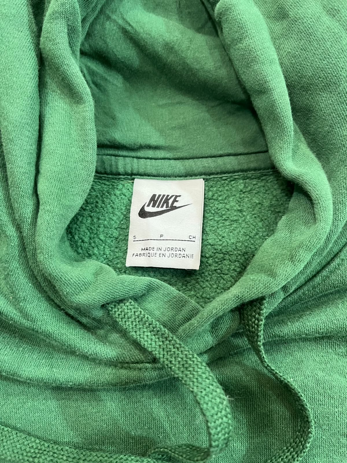 Close-up of a green Nike hoodie showing the inner neck label with the Nike logo and 'MADE IN JORDAN' text, along with green drawstrings and hood fabric.