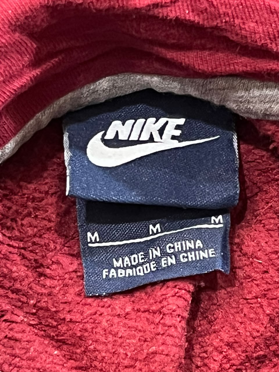 Close-up of a Nike garment label sewn onto a red textured fabric, with the Nike swoosh logo visible on a dark blue tag.
