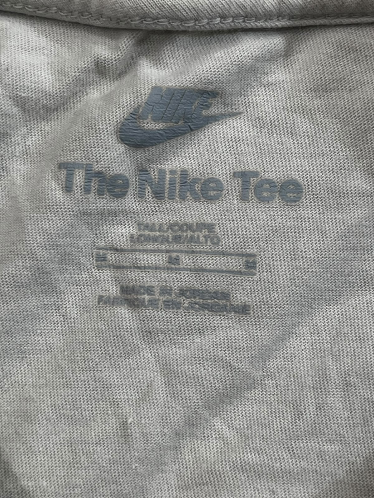 Close-up of the inside neck label of a light gray heather t-shirt, featuring a blue-gray Nike swoosh and 'The Nike Tee' text.