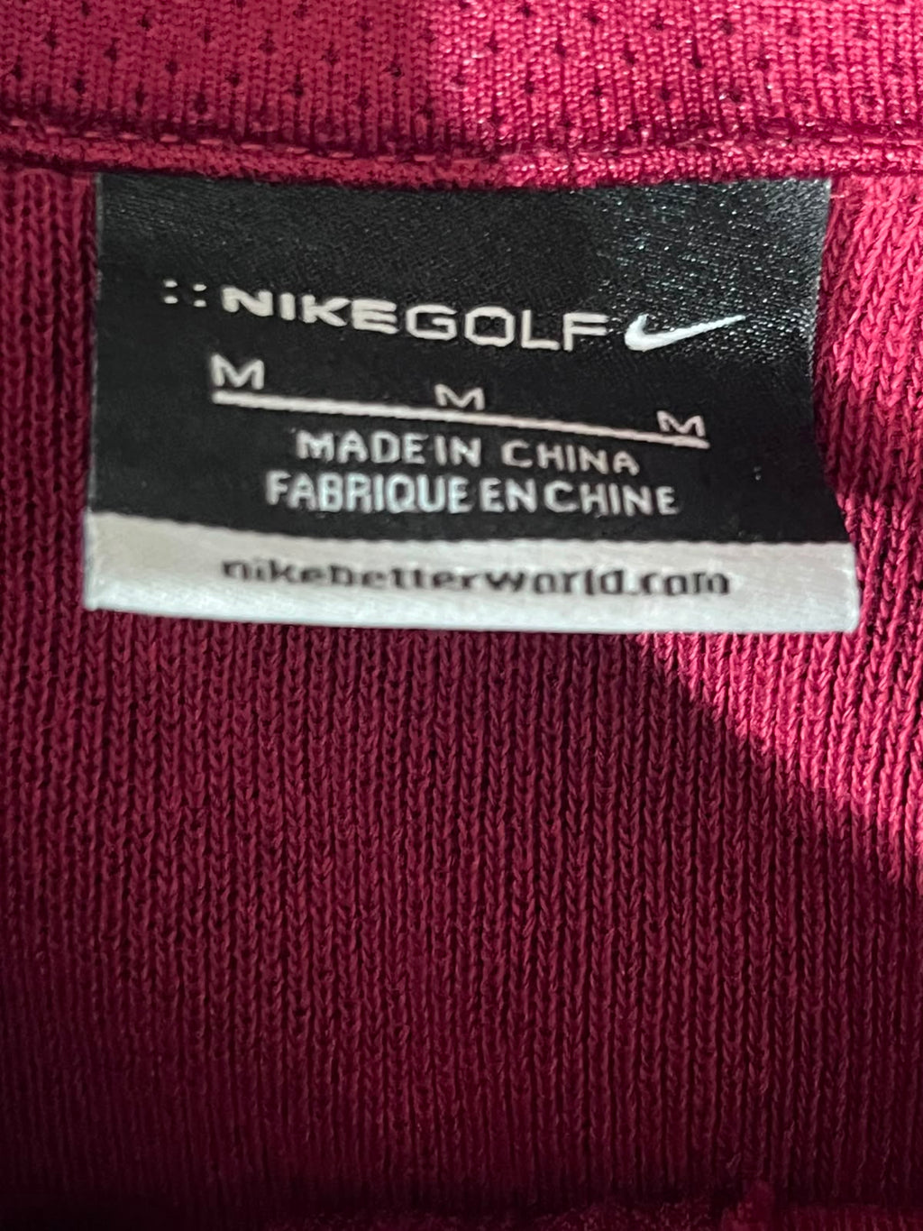 Close-up of a Nike Golf garment label sewn onto a dark pink ribbed knit fabric, showing the Nike Golf branding and size information.
