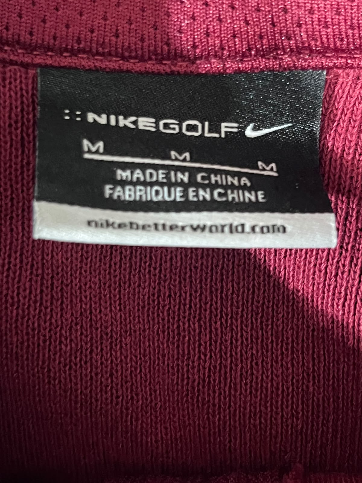 Close-up of a Nike Golf garment label sewn onto a dark pink ribbed knit fabric, showing the Nike Golf branding and size information.