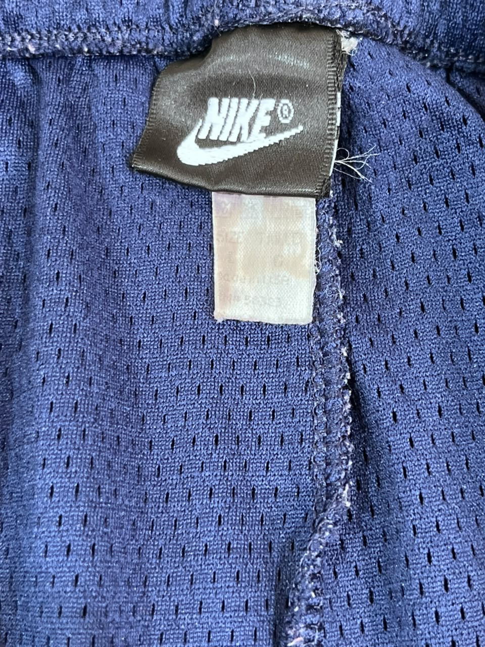 Close-up of a Nike garment label sewn onto a blue mesh fabric, showing the white Nike logo on a dark brown tag.