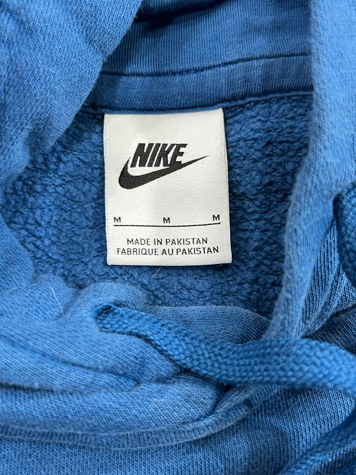 Close-up of a blue Nike hoodie showing the inner neck label with the black Nike logo and text 'MADE IN PAKISTAN' on a white tag.