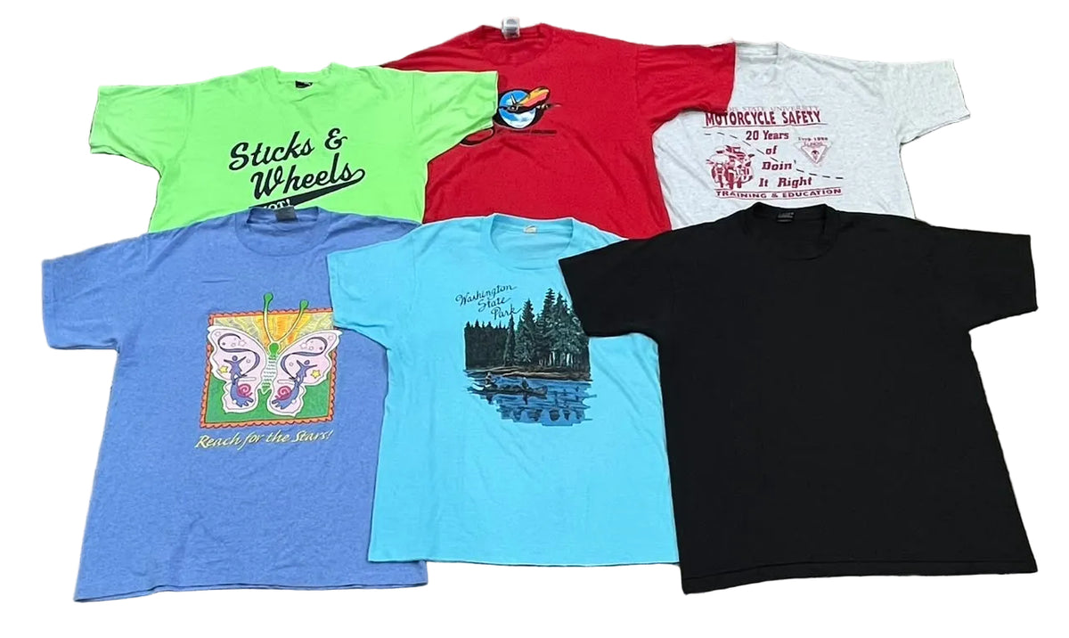 A collection of six vintage graphic t-shirts in various colors (green, red, white, blue, light blue, and black) arranged on a speckled gray floor. Each shirt features a different graphic design, including 'Slicks & Wheels' on the lime green shirt, a butterfly on the blue shirt, and a forest scene on the light blue shirt.