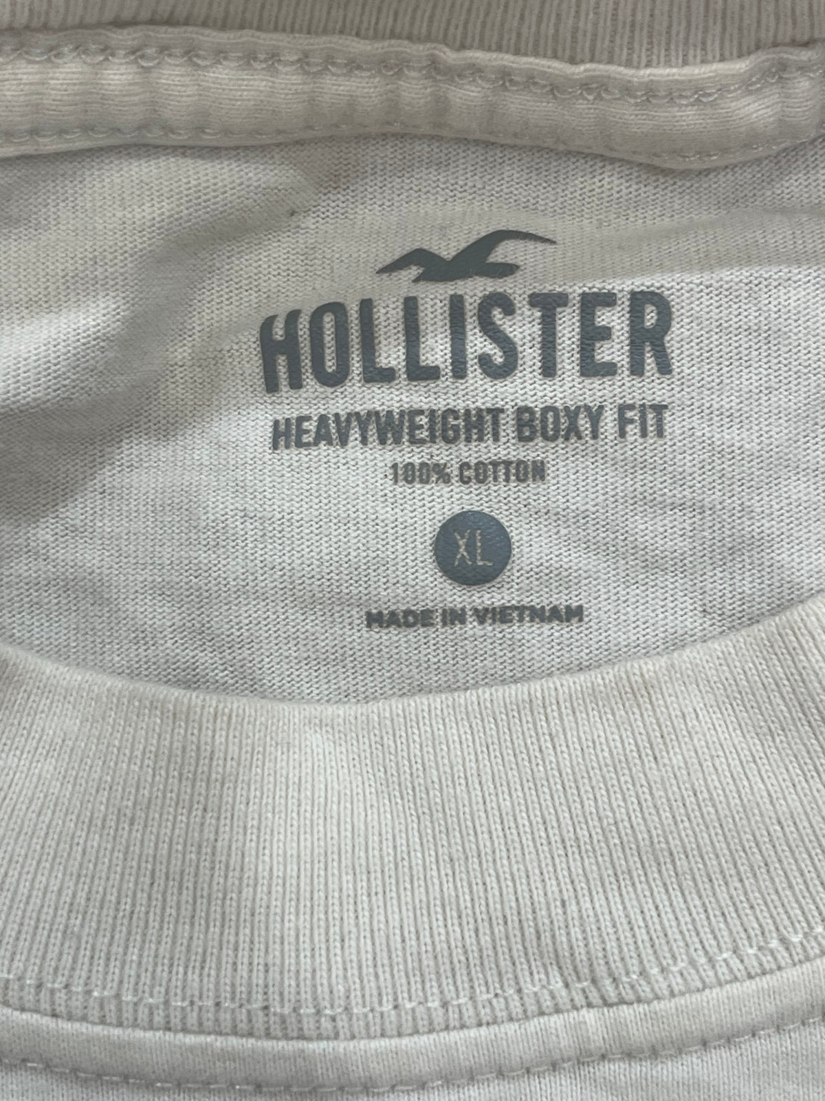 Close-up of the inside neck label of a light gray Hollister t-shirt, showing the brand name, 'HEAVYWEIGHT BOXY FIT', '100% COTTON', and size 'XL'.