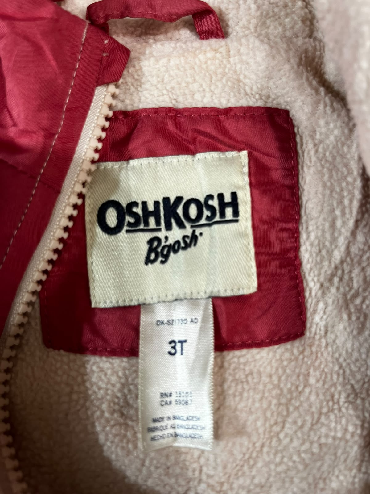 Close-up of a fleece-lined jacket interior showing a cream OshKosh by OshKosh brand label sewn onto a red inner pocket, with a beige sherpa-like exterior visible around it.