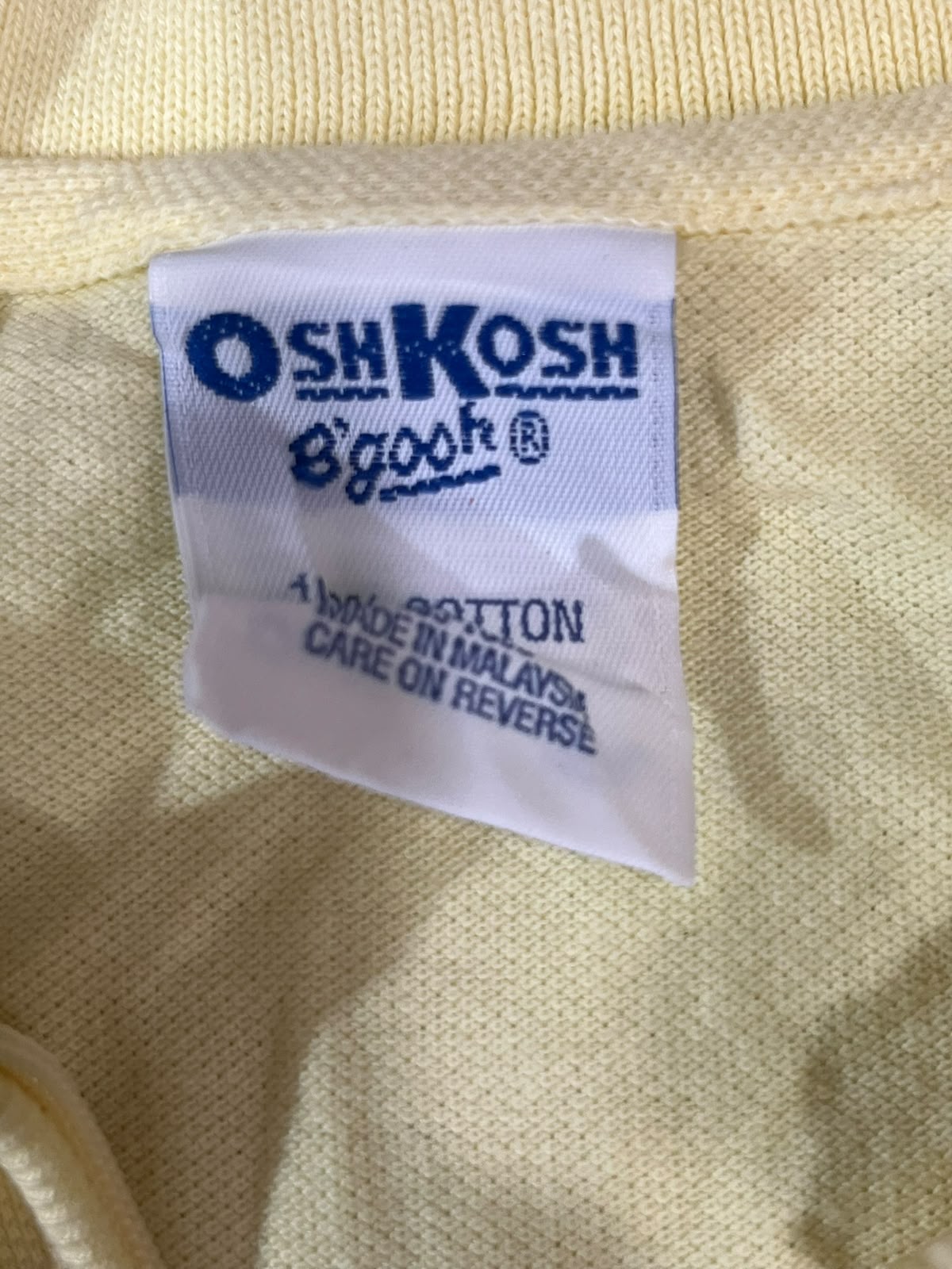 Close-up of a white fabric brand label sewn onto a pale yellow knit garment. The label reads 'OSH KOSH Biggosh' in blue text, with '100% COTTON' and 'MADE IN MALAYSIA CARE ON REVERSE' below.