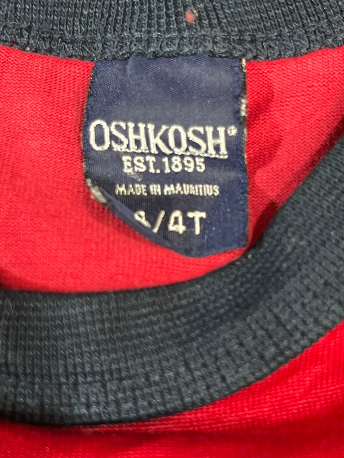 Close-up of a vintage red and black knit top showing the interior neck label which reads 'OSHKOSH EST.1895 MADE IN MAURITIUS' and size '8/4T'.