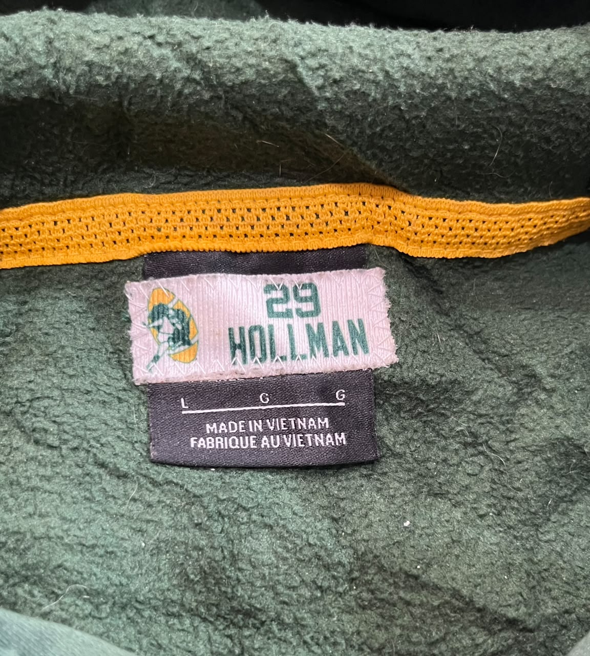 Close-up of a vintage green fleece garment neck label, featuring a pink and black size tag with the name 'HOLLMAN' and number '29'.