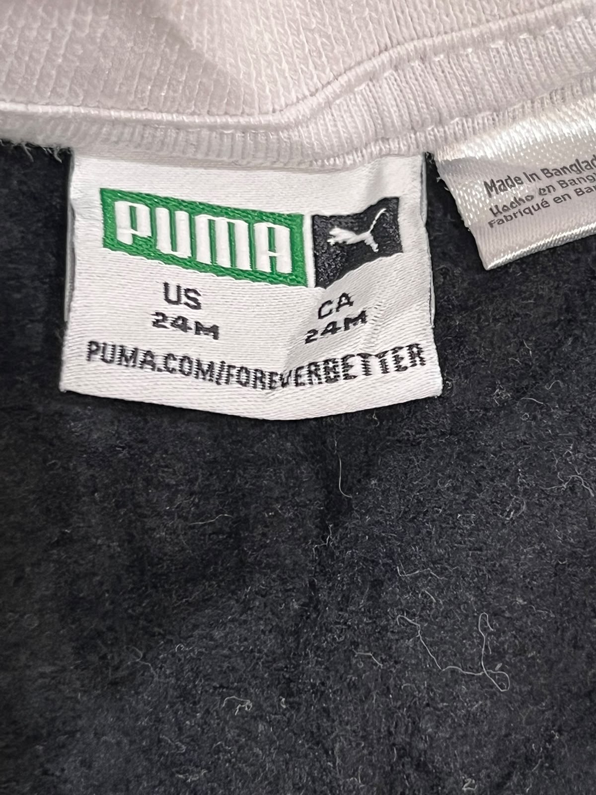 Close-up of a Puma garment label sewn onto a dark gray or black textured fabric, with part of a light-colored ribbed collar visible at the top. The main label reads PUMA with size information and the website PUMA.