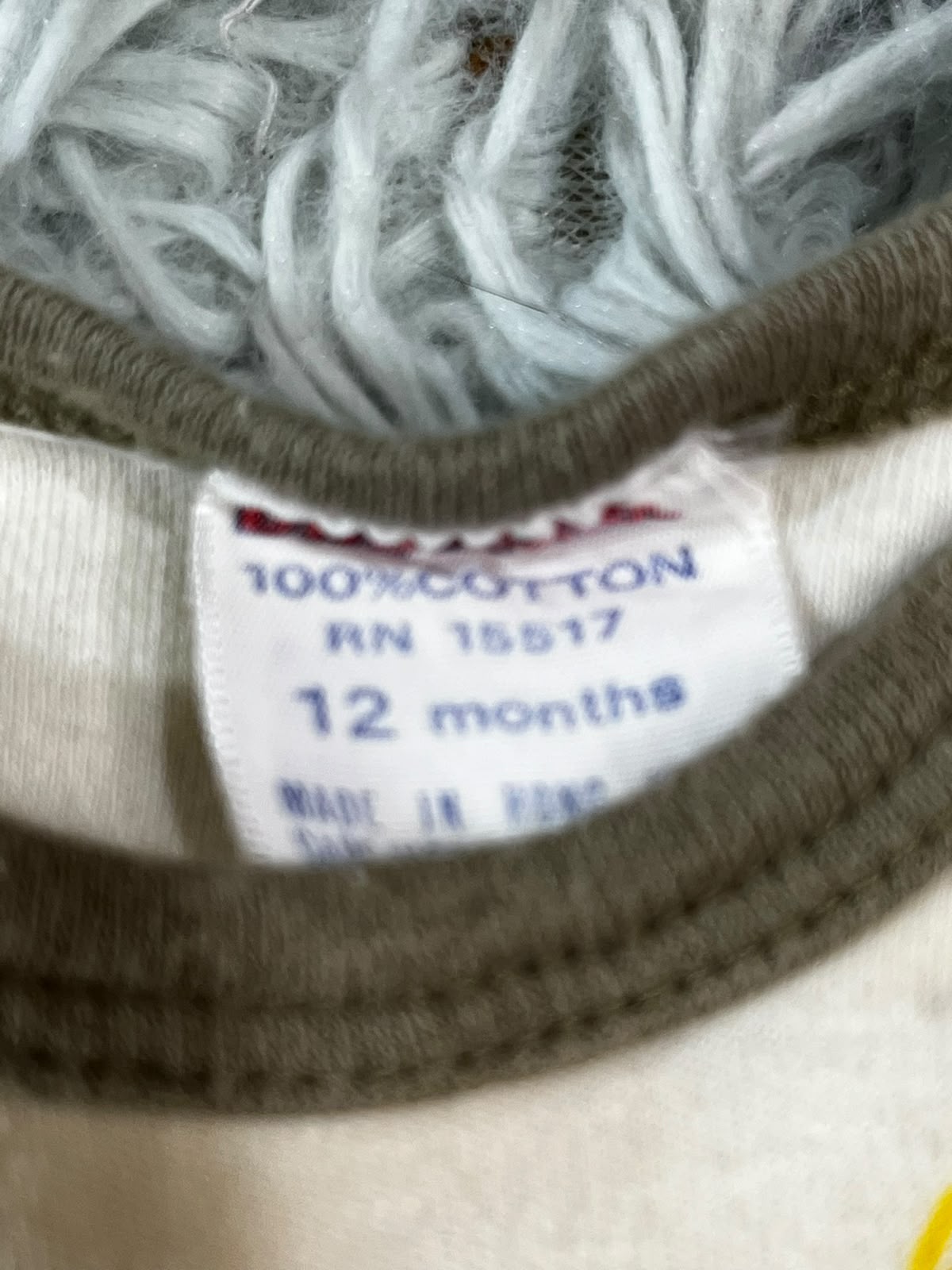 Close-up of a garment neck label reading 'MURPHY' and '100% COTTON RN 15517 12 months', with a brown ribbed collar, off-white fabric, and a pale blue fuzzy textile behind it.