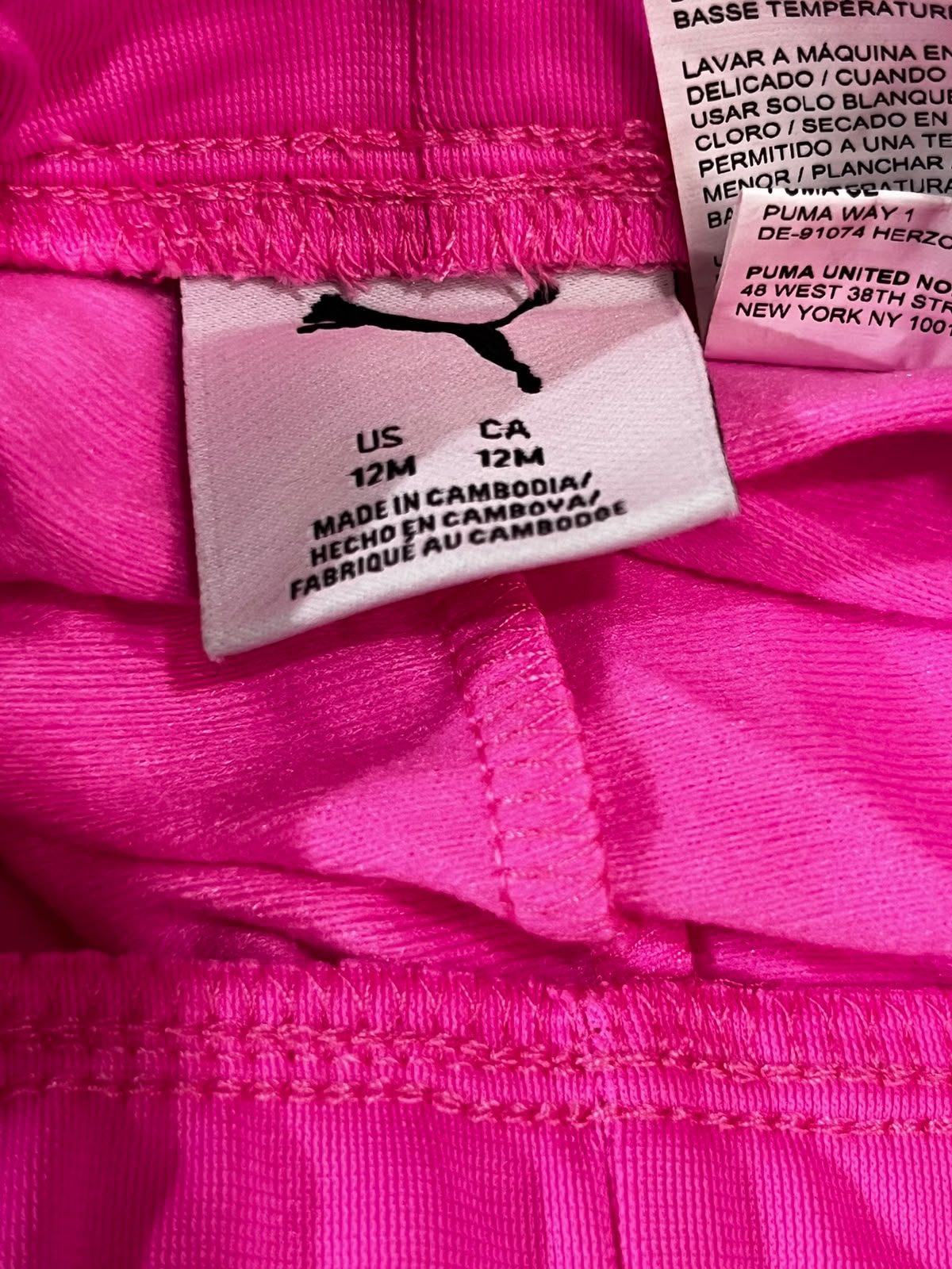Close-up of bright pink fabric with a white Puma size tag showing the Puma cat logo and size information, plus a partial care label.