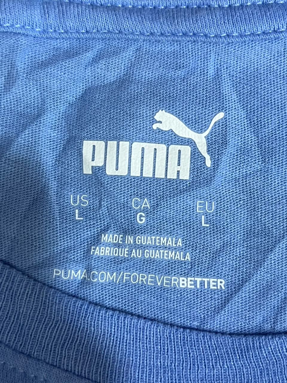 Close-up of a blue Puma garment showing the white Puma logo, size markings (US L, CA G, EU L), 'Made in Guatemala', and 'PUMA.COM/FOREVERBETTER' printed on the inner neck label.