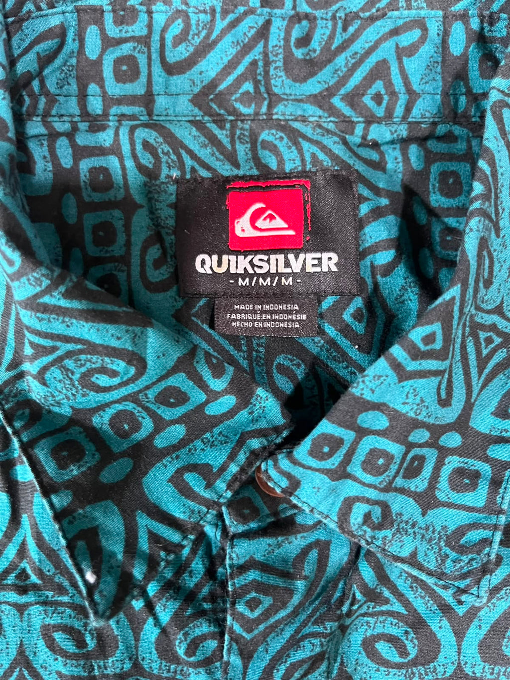 Close-up of a Quiksilver short-sleeve button-up shirt with a black and turquoise geometric tribal pattern, showing the brand label at the collar.
