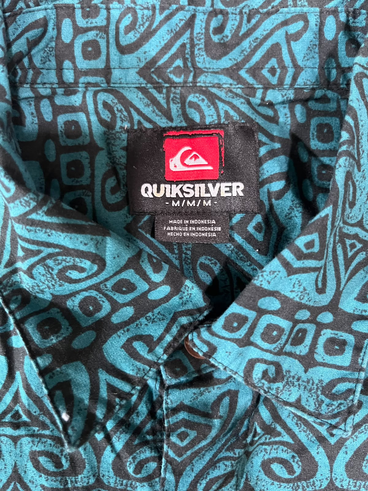 Close-up of a Quiksilver short-sleeve button-up shirt with a black and turquoise geometric tribal pattern, showing the brand label at the collar.