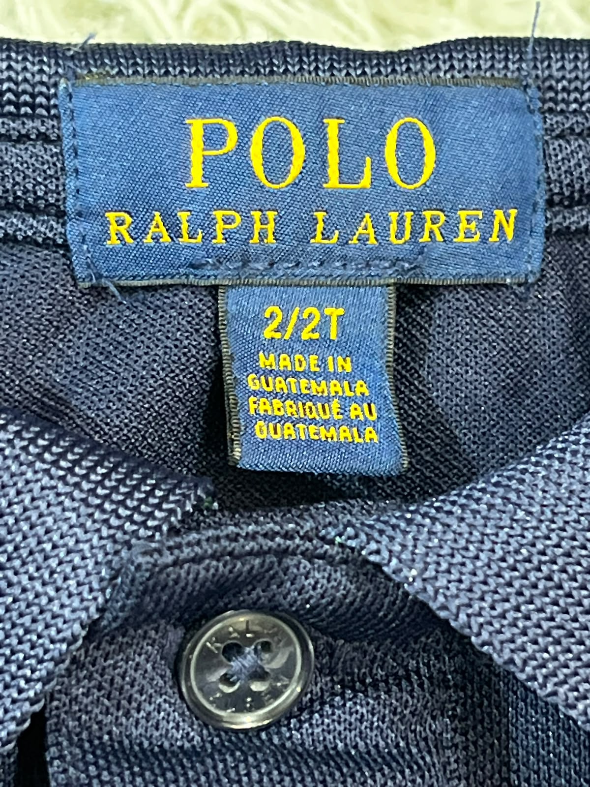 Close-up of a blue Polo Ralph Lauren button-down shirt collar, showing the brand label with yellow text and a button on the front.