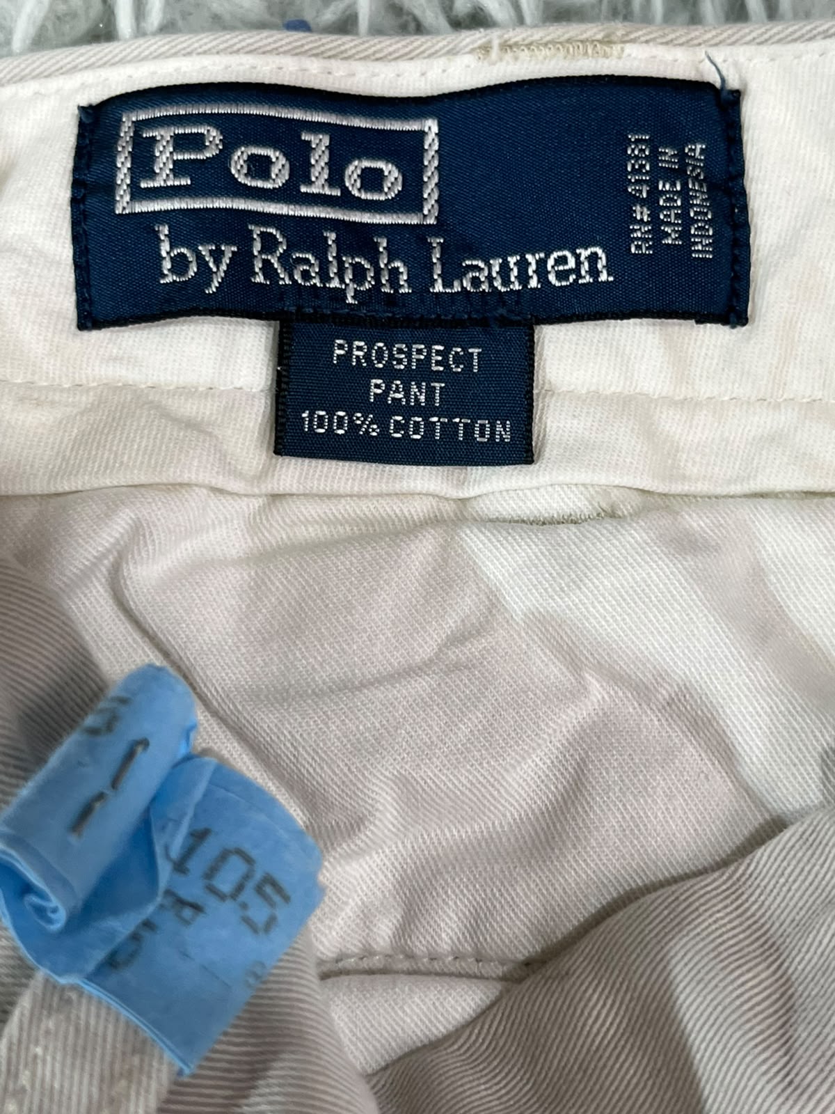 Close-up of a Polo by Ralph Lauren garment label on off-white fabric, with a blue size tag visible in the lower left corner.