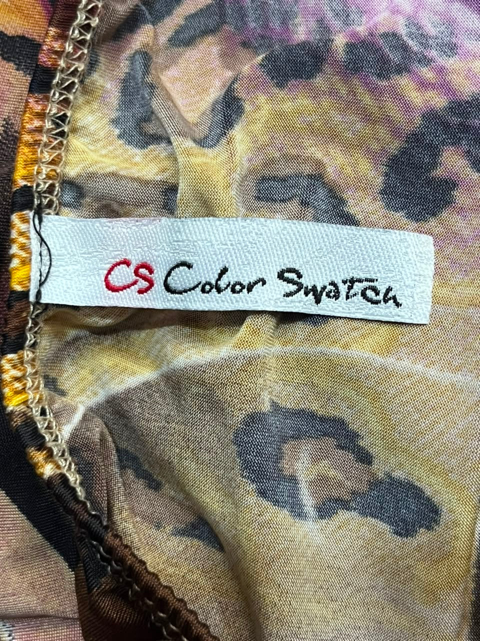 A close-up of a fabric color swatch label reading 'CS Color Swatch' sewn onto a leopard-print garment. The fabric features a beige, brown, and black animal pattern with gold-toned stitching along the edge.