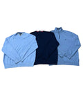 Three long-sleeve quarter-zip knit sweaters a light blue on the left, a dark navy in the center, and a medium blue on the right. Bare feet are visible at the bottom edge, and other folded clothes and shoes appear in the background.