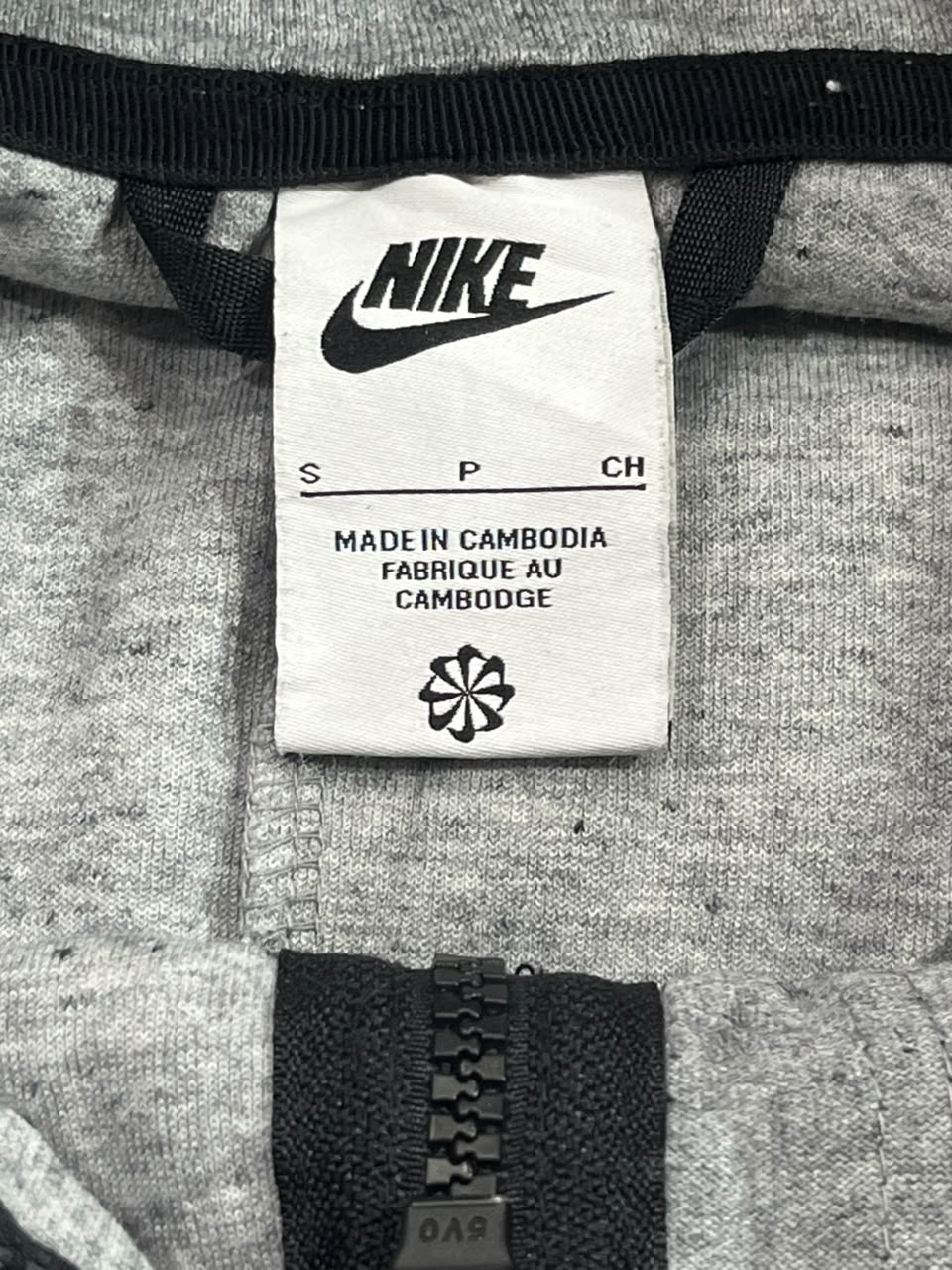 Close-up of a gray heather zippered garment showing the white Nike brand label with black text and logo, along with black zipper trim.