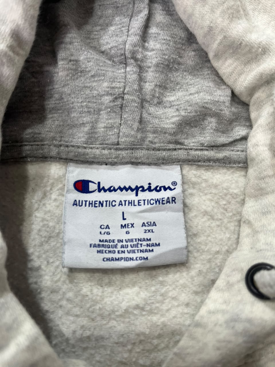 Close-up of a Champion athletic hoodie interior label, showing the Champion brand name and Authentic Athleticwear text on a white tag, with light heather gray fabric surrounding it.