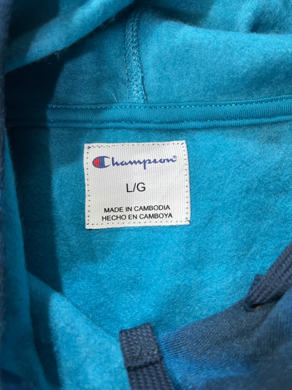 Close-up of a Champion hoodie interior label showing the brand name, size L/G, and Made in Cambodia text, with turquoise blue fabric surrounding the label.