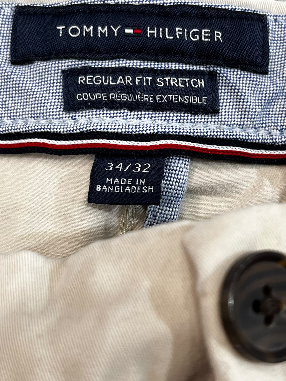 Close-up of Tommy Hilfiger garment labels on a light beige fabric item, including a navy brand label, a stretch label, and a size/origin tag.
