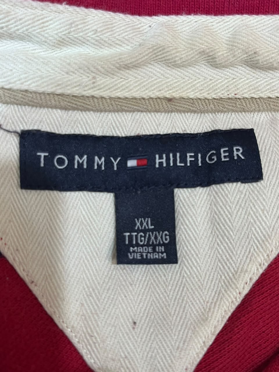 Close-up of a Tommy Hilfiger garment neck label on a cream herringbone fabric, with red fabric visible along the sides. The main label reads TOMMY HILFIGER, and the smaller size tag reads XXL TTG/XXG MADE IN VIETNAM.
