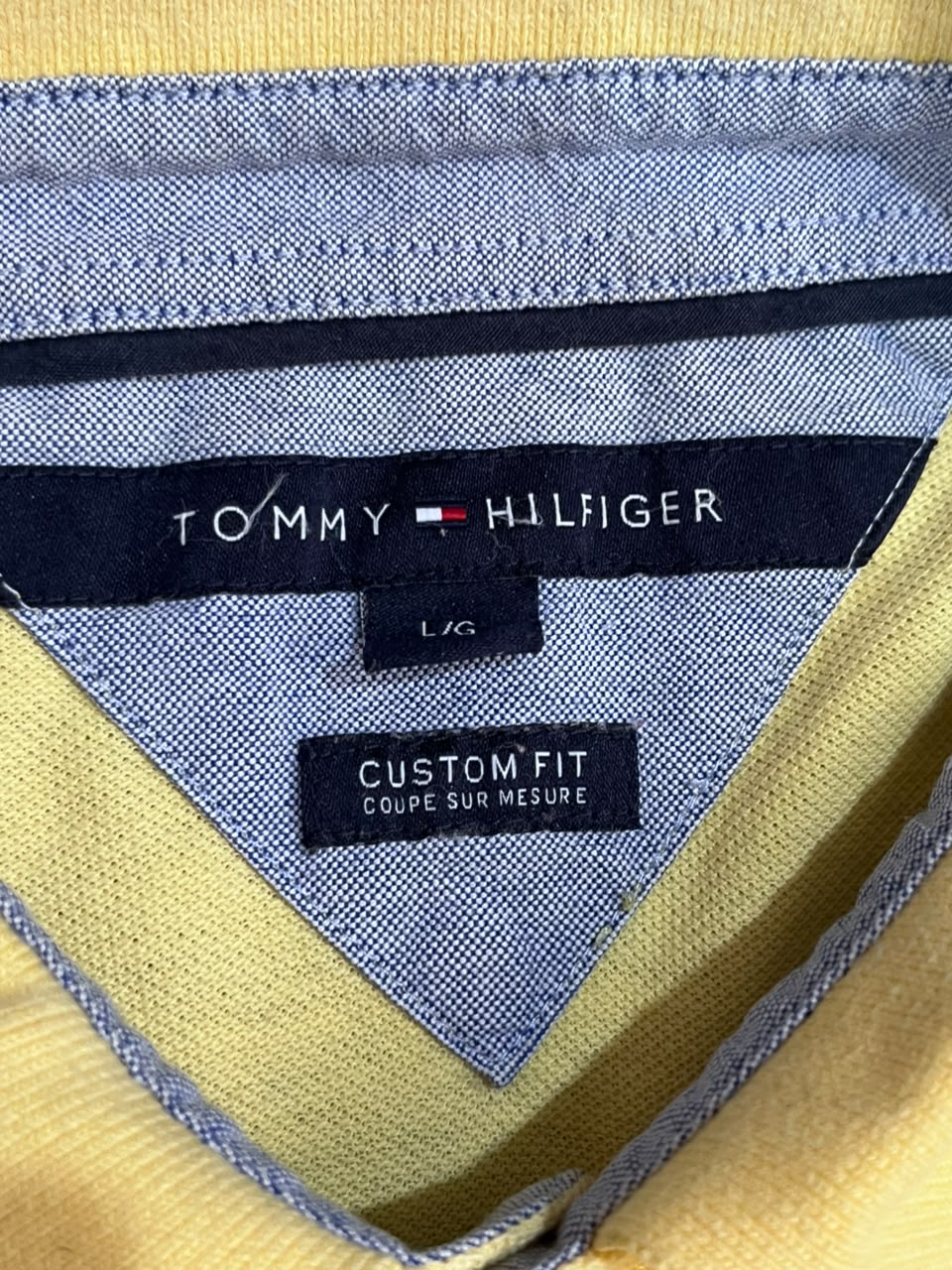 Close-up of the inner neck label of a Tommy Hilfiger garment, showing the brand name, 'L/G' size, and 'CUSTOM FIT COUPE SUR MESURE' on a light blue textured fabric background.