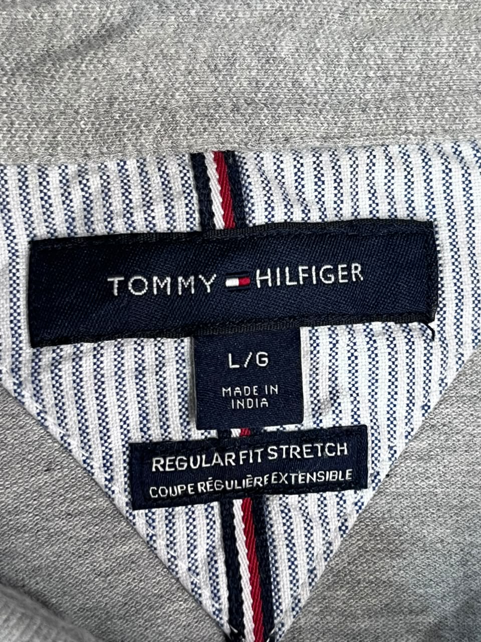 Close-up of a Tommy Hilfiger garment interior label on a gray knit top, showing the brand name, size L/G, made in India, and stretch label details.