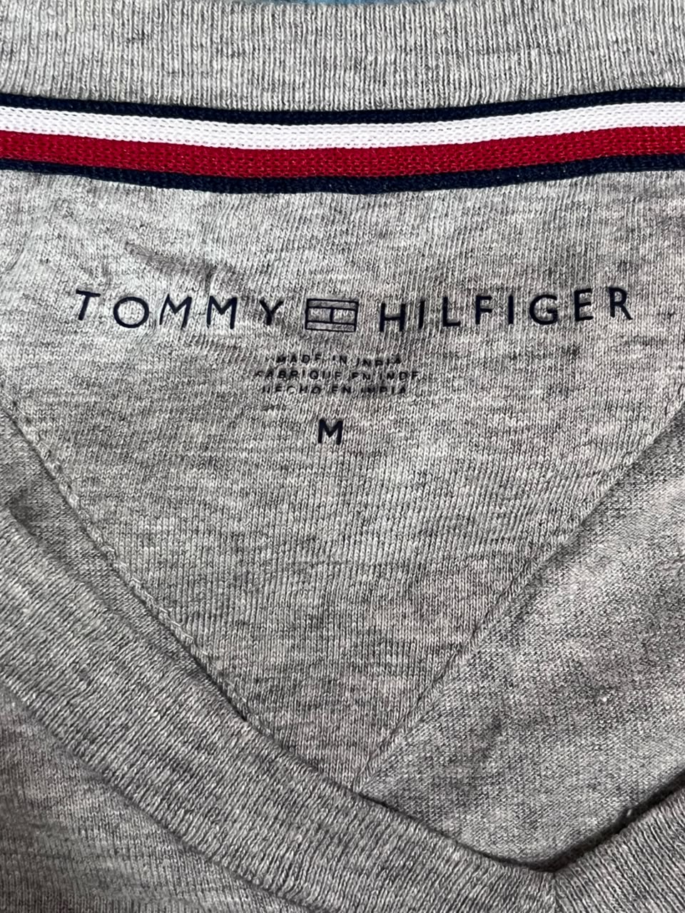 Close-up of a heather gray Tommy Hilfiger knit garment, showing the inner neck label and the signature red, white, and navy striped collar trim.