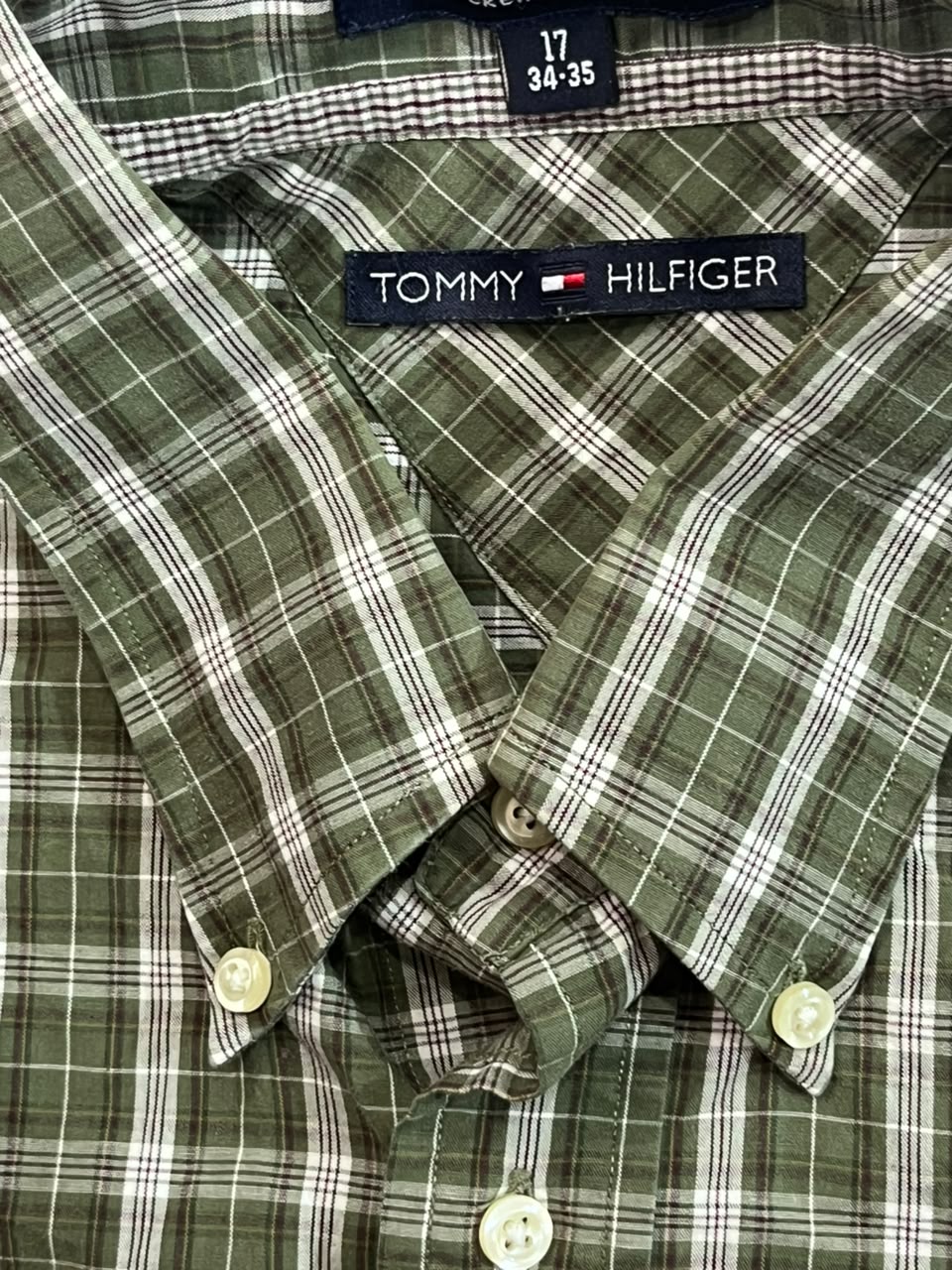 Close-up of a Tommy Hilfiger plaid button-down shirt in olive green, white, and black, showing the collar, brand label, and buttons.