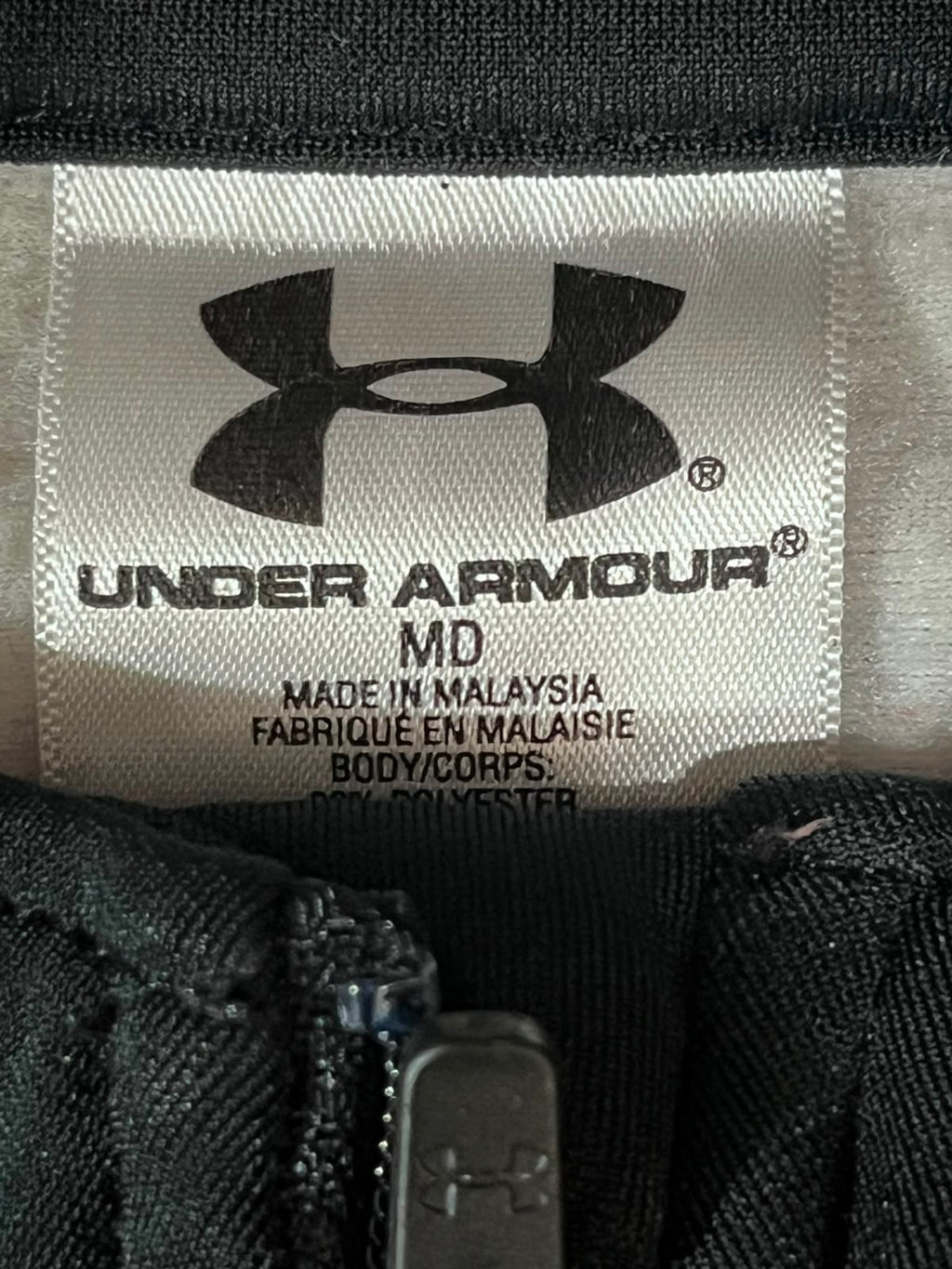 Close-up of an Under Armour garment label showing the black Under Armour logo and 'UNDER ARMOUR' text on a light fabric tag, with black zipper trim visible around it.