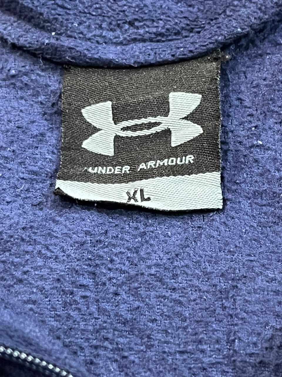 Close-up of a Under Armour garment label on a textured blue fleece or sweatshirt fabric, showing the Under Armour logo and size XL.