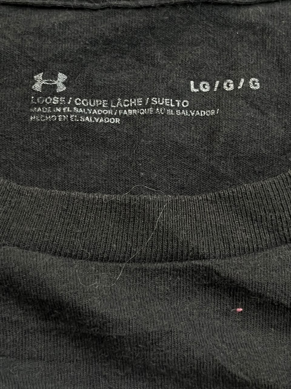 Close-up of the inside collar of a dark charcoal gray Under Armour t-shirt, showing the white printed Under Armour logo, size information 'LG/G/G', and country of origin text.