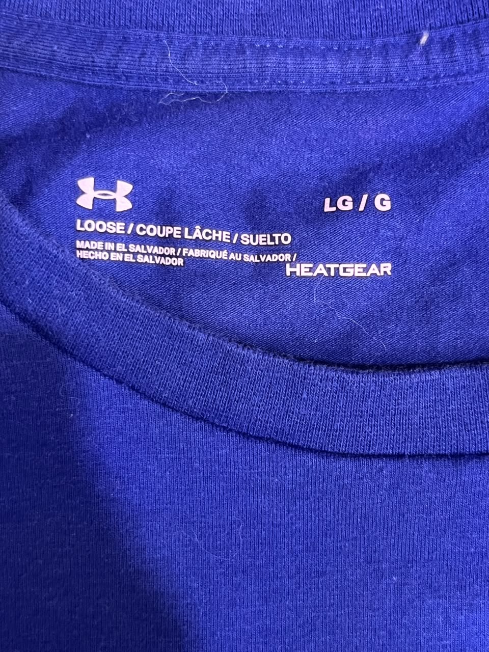 Close-up of the inside collar of a royal blue Under Armour Heatgear T-shirt, showing the white printed Under Armour logo, size information (LG / G), and manufacturing details.